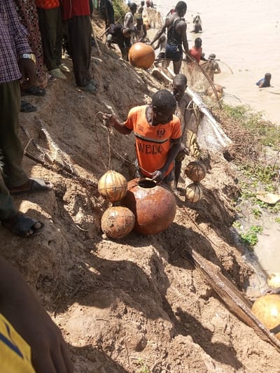 The image displays a scene on a sloping, light brown sandy bank adjacent to a murky body of water in Kokani North, Nigeria. In the central foreground, a dark-skinned male wearing an orange t-shirt with "WELL" printed on it holds a rope connected to a light brown spherical object, which appears to be a gourd or a float. This individual is positioned near a large, reddish-brown clay pot and two additional similar spherical objects resting on the sand. Behind him and to the right, another individual is partially visible.

Further up the bank and extending into the background, multiple other dark-skinned individuals are present. Some are standing on the eroded sandy incline, while others are partially submerged in the light brown water, engaged in activities involving fishing nets or traps. A large, conical-shaped fishing net structure is visible on the bank in the mid-ground. Additional light brown spherical objects, consistent with gourds or floats, are scattered on the bank near the water's edge. The water itself contains sparse green aquatic vegetation along the bank. The lighting suggests bright daylight, casting distinct shadows. Individuals are dressed in casual attire, including t-shirts, pants, and sandals; some in the water are shirtless.