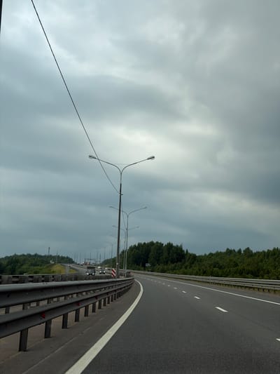 The image shows a highway in Korabsel'ki, Russia. The highway is empty except for a few cars in the distance. The sky is overcast, and the weather is cloudy. There are trees on the side of the highway, and there are streetlights on the side of the road. The image is taken from the perspective of someone driving on the highway, looking forward. The road is asphalt and has white lines marking the lanes. The highway guardrail is visible on the right side of the image. The trees are green and lush, suggesting a temperate climate. The lighting is dim, indicating it's likely daytime but not a bright, sunny day. No people are visible in the image.