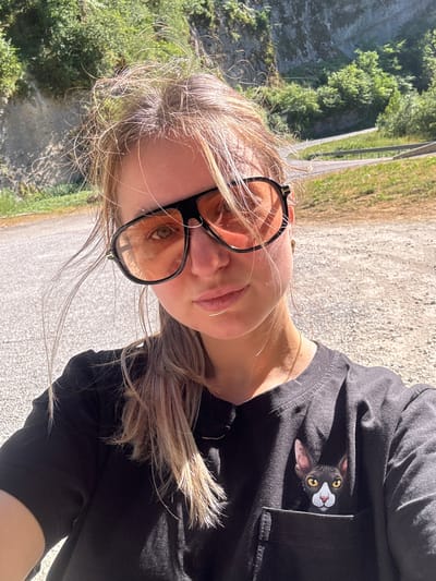 A young woman with blonde hair and sunglasses is taking a selfie outdoors. She is wearing a black t-shirt with a printed image of a cat peeking out of a pocket. The background shows a steep, rocky cliff face covered in greenery, with a winding road visible in the distance. The lighting suggests it is a sunny day. The image does not contain any information that allows for identifying the location in the city of L'Essard, France.