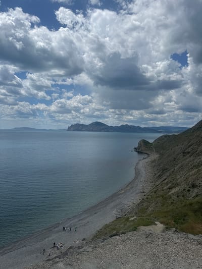 The image shows a coastal scene in Орджоникидзевский поселковый совет, Ukraine. The sky is partially covered with white and gray clouds. The sea is calm and blue-gray. A rocky cliff dominates the right side of the image, sloping down to a pebble beach. Some people are on the beach. A metal structure is located on the beach near the cliff. Mountains are visible in the distance, partially obscured by haze.