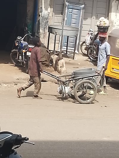 The image captures a sunlit street scene in Kofar Mazugal, Nigeria. In the center, a young male, wearing a dark reddish-brown long-sleeved shirt and light brown trousers, pushes a two-wheeled cart equipped with a large, metallic, circular basin. His right foot is bare, and his left foot wears a light-colored slipper. To his immediate right, an adult male stands, wearing a light blue tunic and light-colored slippers, balancing a black bucket filled with plastic-wrapped items on his head.
In the background, a dark building entrance is visible on the left, next to a parked blue and white motorcycle. Further right, a weathered light-colored building wall features a prominent light blue metal door leaning against it, and a metallic circular table-like structure on a stand. Towards the far right, a partial view of a yellow auto-rickshaw and another motorcycle are present. The ground is a mix of dirt and asphalt. The front section of a black scooter is visible in the lower left foreground.