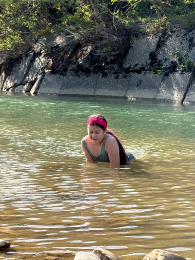 The media file shows a person partially submerged in shallow, murky water. The person appears to be a young female wearing a green swimsuit and a pink headband. Her eyes are closed, and she is smiling, suggesting a relaxed or happy mood. The water is clear enough to see the rippled sandy bottom.
The scene is set in a natural outdoor environment, likely a river or a lake, given the presence of rocks and vegetation along the bank. The lighting suggests it is daytime, with sunlight casting reflections on the water's surface. The water is a greenish-brown color, and the sunlight creates bright, wavy patterns on it.
To the left of the image, there is a rocky bank with some vegetation, including trees and bushes. The rocks are grey and appear somewhat rough, with some moss or dark growth on them. The overall setting suggests a peaceful, natural location, possibly in Krasnoye, Russia, as suggested by the location context. There is no visible text in the media file.