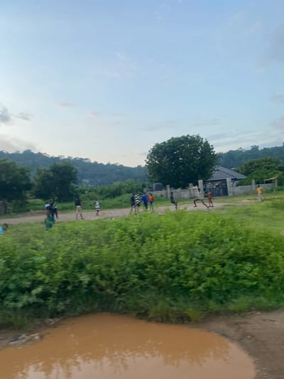 This image depicts a semi-rural landscape in Uffah, Nigeria, under a pale blue sky with minimal cloud cover. In the far background, verdant hills and mountains rise, densely covered in trees.
The midground features an unpaved dirt path that horizontally traverses the scene. Along this path, a scattered group of approximately twelve individuals, predominantly male and appearing to be young adults or adolescents, are present. On the left side of the path, a cluster of individuals, mostly in dark or light shirts and shorts, are standing or casually moving. On the right side, another group of individuals exhibits more dynamic postures; one person in a dark top and light shorts is captured mid-air or in a wide-legged stance, another in an orange top and dark shorts is also in a dynamic, possibly kicking or leaping, pose. A person in a yellow top stands near a concrete perimeter wall on the far right.
Behind the path on the right, a single-story building with a dark, sloping roof and light-colored walls is partially visible, enclosed by a gray concrete wall or fence that features a dark metal gate. A large, dark, cylindrical object, possibly a water storage tank, is situated to the left of the building. Large, dense green trees are interspersed throughout the midground, both behind the path and near the building.
The foreground is dominated by a large, irregularly shaped puddle of muddy, reddish-brown water, which occupies the lower center and right portions of the frame and reflects the sky. Abundant green foliage, consisting of bushes and tall grass, fills the lower left and central foreground, extending towards the unpaved path. The overall impression is an outdoor setting with human activity amidst natural and constructed elements.