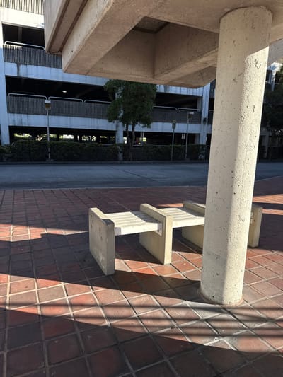 A low-angle shot shows a concrete architectural setting. A large, cylindrical concrete beam extends horizontally above. To the right, the beam casts a shadow on a red brick wall with protruding concrete steps. In the background, a parking structure is visible, with trees lining the side. The image is rotated sideways.