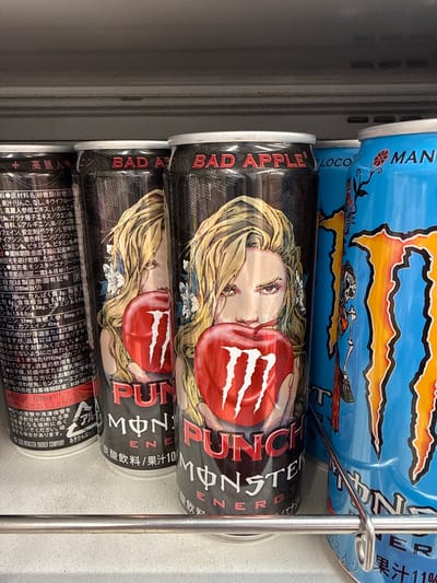 The image shows several cans of Monster Energy drink on a shelf. The cans are predominantly black and blue, with the Monster Energy logo visible on each. Two cans in the foreground have an illustration of a woman holding a red apple with the Monster logo on it. Text on the cans indicates they are "BAD APPLE PUNCH" and "MONSTER ENERGY." To the right, a blue can also displays the Monster Energy logo and some Asian script. The environment appears to be a retail setting, likely a convenience store or supermarket, with a grey shelf above and a metal rack supporting the cans in the foreground. The background is blurred, suggesting a shallow depth of field. A small portion of text on the leftmost can, when viewed closely, indicates a "2005 MONSTER ENERGY COMPANY" copyright and mentions "ADACHI" and "TOKYO."