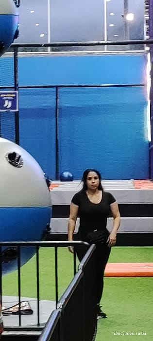 A woman in black clothing is standing in a room with colorful walls and equipment. She appears to be in the middle of an athletic activity, possibly stretching or preparing to use the equipment. The room has blue and green padded walls, with metal bars and a large white and blue inflatable structure visible. The overall impression is that of an indoor recreational or training facility.