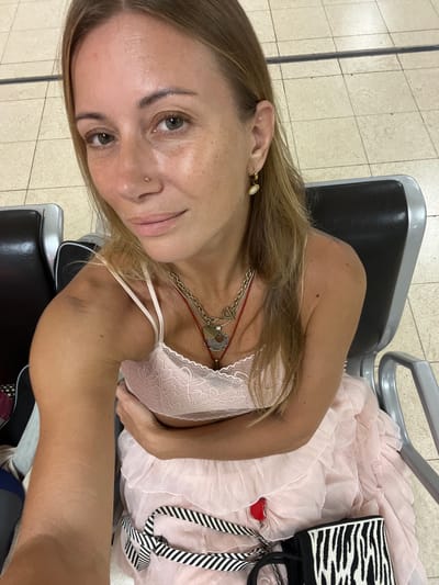 A woman with blonde hair and green eyes is sitting in a waiting area, likely an airport lounge, in Katunayake, Sri Lanka. She is wearing a light pink lace camisole and a layered pink dress. A red item, possibly a charm or tag, is attached to her dress. She is also wearing a necklace with multiple pendants and earrings. A bag with a black and white zebra print is on her lap, secured by a striped strap. The environment appears to be an indoor seating area with tiled flooring and dark, upholstered chairs.