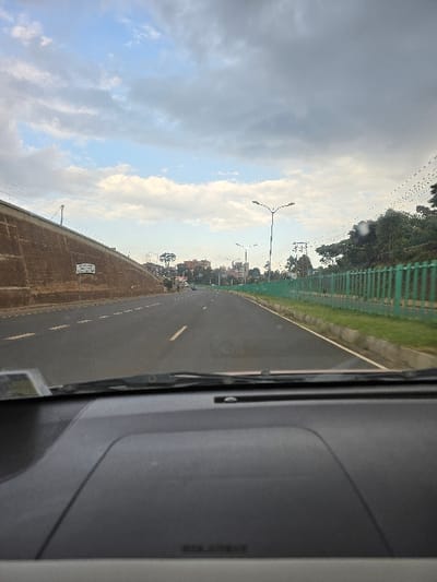 This media file captures a view from inside a vehicle, looking forward through the windshield at a road scene in Wangige, Kenya. The paved road, marked with dashed white lines, stretches into the distance, appearing to curve slightly.

To the right of the road, a green metal mesh fence runs parallel, beyond which are numerous green trees and foliage. Further in the background, several streetlights with dual arms are visible, along with indistinct buildings and structures, suggesting an urban or semi-urban setting. Overhead, utility lines traverse the sky.

On the left side of the road, a low, tan-colored wall, possibly constructed from brick or stone, extends from the foreground. A rectangular white sign is affixed to this wall; while most of the dark text is blurred, the word "MWEMBE" is discernible on the top line.

The sky is largely overcast, featuring a mix of grey and white clouds, indicating a daytime scene with diffused lighting, likely late morning or afternoon. The weather appears calm. A portion of the vehicle's dark-colored dashboard and interior trim is visible on the right side of the frame. No individuals are present in the image.