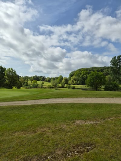 The image presents a landscape in Lenningen, Luxembourg, likely a golf course or park. A dirt path runs vertically through the right side of the image. To the left of the path is a well-maintained green lawn, suggestive of a fairway. Beyond the lawn, trees and foliage are present, forming a natural boundary. The sky occupies the left portion of the image, filled with scattered white clouds against a blue backdrop. There are no visible people, subjects, or explicit actions in this scene.