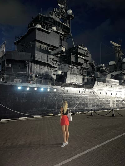 A woman with blonde hair, wearing a black top and a red skirt, stands facing away from the camera on a brick-paved area. She is looking at a large, dark-colored ship docked at night. The ship has several levels of structure and appears to be a museum or historical exhibit. Lights are visible along the side of the ship, and the number "103" is painted on its hull. The sky is dark with some clouds. The scene is set in the city of Verkhnyaya Stavropol'ka, Russia.