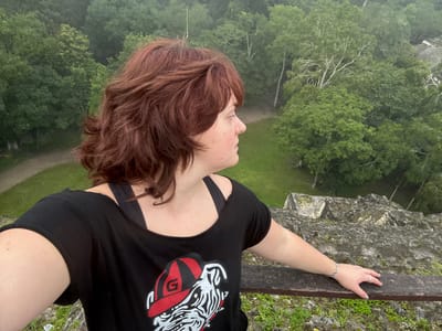 An adult female is depicted in an elevated, outdoor setting, likely atop an ancient stone pyramid or temple within Tikal National Park, near Flores, Guatemala. The subject, viewed from the upper torso, has reddish-brown, wavy hair and wears a black t-shirt with a white and black graphic of an animal wearing a red cap marked with a white "G". A black undergarment is visible beneath the t-shirt. She wears a silver-colored bracelet on her left wrist and a silver-colored ring on her left ring finger. Her right forearm rests on a weathered wooden railing situated at the edge of a gray, moss-covered stone structure. The subject's head is turned to the right, observing the distant landscape. The background reveals a vast expanse of dense green jungle canopy, with a winding dirt path, grassy clearings, and the partially visible upper sections of other stone structures integrated into the forest below. The sky overhead appears overcast or hazy.