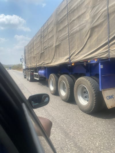 The image depicts a highway scene in Ninzo West, Nigeria, from the interior perspective of a moving vehicle. A large white articulated truck with a blue chassis and a tan tarpaulin-covered cargo is prominently featured, traveling parallel to the observer's vehicle. The truck's trailer has three visible axles with dual wheels, and a mudflap displaying "CIMC" is visible on the rear-right side. Blue air tanks are mounted beneath the trailer. The road is paved asphalt, and two additional vehicles are visible in the distance ahead on the same road. The sky is bright blue with scattered white clouds, and a distant treeline and hills are visible on the horizon. In the immediate foreground, a dark-skinned human arm is resting on the window sill of the observer's vehicle, and a reflection of a person's head/upper body is partially visible in the vehicle's side mirror.