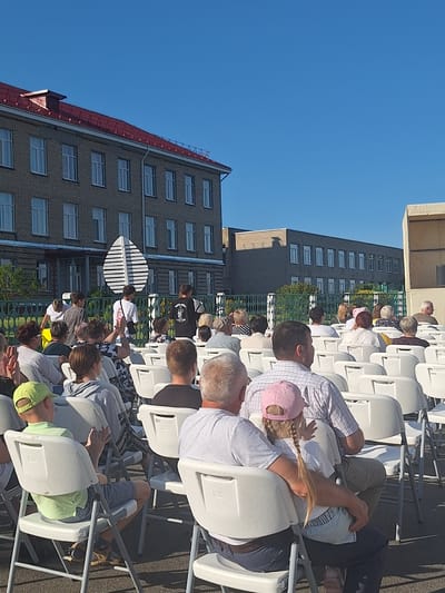 A group of people are seated in white plastic chairs outdoors, facing a stage area that is partially visible on the right side of the frame. The setting appears to be a school or community event, with a large brick building with a red roof in the background. The sky is clear and blue, suggesting it is a sunny day. Some individuals are looking towards the stage, while others are interacting with each other. The overall atmosphere seems relaxed and social.