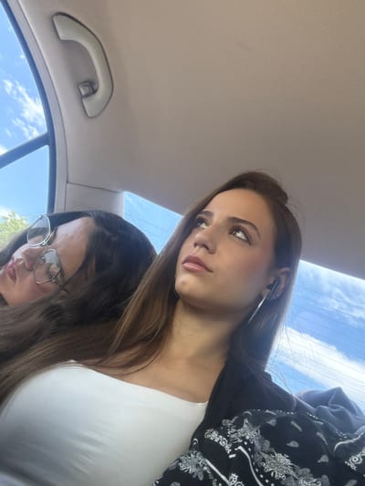 The image depicts two young women inside a car, potentially in motion, based on the background view of a partly cloudy sky through the car window. The woman in the foreground is fair-skinned with long brown hair, wearing a white top and a jacket with a black and white paisley print. She has large hoop earrings and appears to be looking downward and to the left. The second woman, positioned slightly behind and below, also has long brown hair and wears glasses. A car grab handle is visible in the upper left corner of the image. The location is not discernible as Gorni Bogrov, Bulgaria.