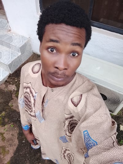 The image is a selfie taken outdoors in Orodo, Nigeria. The subject is a young Black male with dark, curly hair, looking directly at the camera with wide eyes and a neutral expression. He is wearing a tan, patterned long-sleeved garment with blue accents. His left hand is visible, holding a dark-colored smartphone.

The background shows a light-colored, textured wall on the left, with a set of tiled steps descending. To the right of the subject, a white outdoor air conditioning unit is visible. The ground beneath the subject and in the foreground is dark and appears to be a mix of soil and some moss or vegetation. The lighting suggests it is daytime.