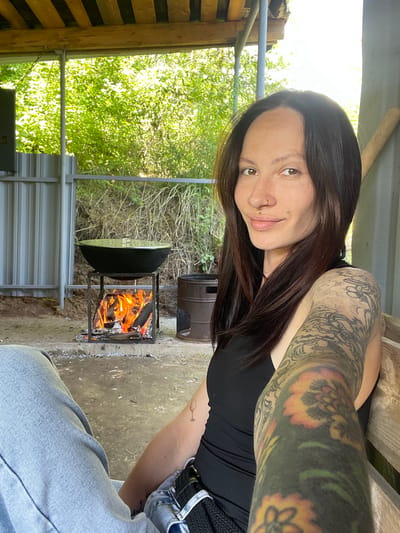 A woman with dark hair and visible tattoos on her left arm poses for a selfie. She is wearing a black tank top. She is situated near a makeshift outdoor cooking area with a metal frame holding burning wood. A large black pan sits above the fire. The environment consists of a simple structure with wooden supports and corrugated metal sheeting, possibly in a rural or semi-rural setting. The background reveals green foliage suggesting a wooded area. The location may be in Doma 45 Kilometr, Russia.