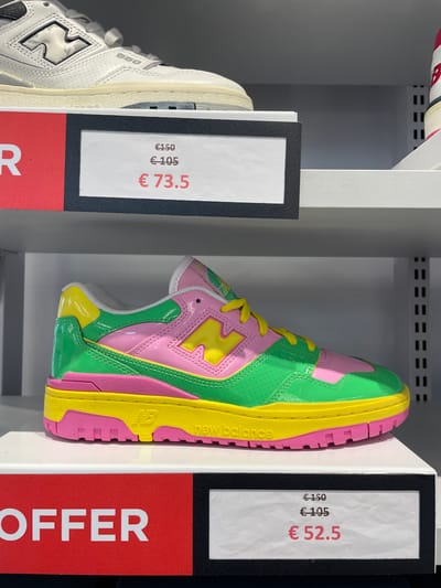 The image captures a display of New Balance 550 sneakers on a shelf, likely in a retail store, as indicated by the "OFFER" signs and price tags. Two different colorways of the shoe are visible. On the left, a white and grey sneaker is displayed with a price tag of €73.5. To the right, a vibrant multicolored sneaker (green, pink, and yellow) is displayed with a price tag of €52.5.

While the location information "Marcianise, Italy" is provided, there's nothing in the image itself that definitively confirms this location.