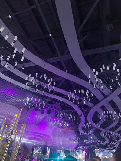 This media file captures a visually striking event, likely a performance or grand celebration, taking place indoors at night. The scene is dominated by a dramatic and intricate lighting design. Hanging from the ceiling are numerous modern chandeliers, each featuring multiple white cylindrical lights arranged in a circular, almost radial pattern. These chandeliers are suspended against a backdrop of flowing, wavy structures in shades of grey and deep purple, suggesting a theatrical or concert setting.

The lower portion of the image reveals a vibrant, pulsating purple light emanating from a large installation that resembles cascading fabric or perhaps a water feature illuminated by stage lights. This light creates a luminous, ethereal atmosphere. Scattered throughout this illuminated area are delicate, cylindrical lights with a soft yellow glow, possibly mimicking candlelight or decorative elements.

On the right side of the image, a group of people are visible, partially obscured by the lighting and structural elements. They appear to be spectators, observing the spectacle before them. Their presence suggests the event is ongoing and attended. The overall mood is one of awe and wonder, amplified by the dramatic lighting and the scale of the setup.

Visible text on what appears to be a banner or screen in the background reads "ORCHESTP" and "BY ADANA COMP," offering a potential clue to the event's organizer or theme. The time of day is clearly night, and the indoor setting eliminates any weather considerations. The location context of Mkhchyan, Armenia, is provided externally.