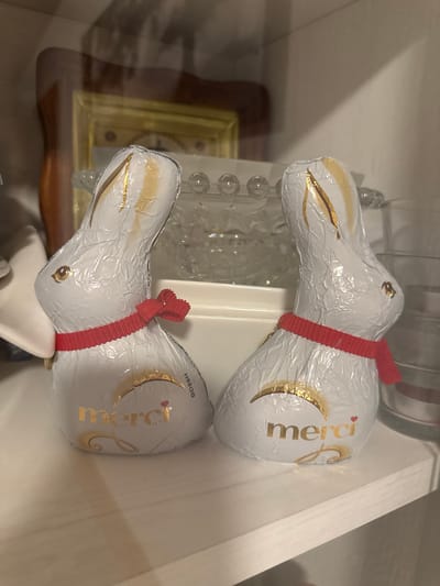 This media file shows two foil-wrapped chocolate easter bunnies. The bunnies are white with gold accents and red ribbon ties. The word "merci" is printed in gold on the body of each bunny. The bunnies appear to be sitting on a shelf or in a display case, with a clock and other decorative items visible in the background. The lighting suggests an indoor setting. The image is oriented vertically, with the bunnies positioned one above the other.