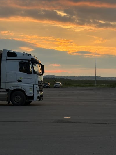 A white semi-truck is parked on an asphalt lot. The truck is in the foreground, taking up the left side of the frame. The truck has "ТЕХНОЛОГИЯ ДВИЖЕНИЯ" written on its side in green and white lettering. The truck is facing towards the right of the frame. In the background, there is a line of trees and hills. The sky is a dramatic mix of orange, yellow, and grey clouds, indicating sunset or sunrise. A single lamppost stands in the background, to the left of the truck. Two smaller vehicles, possibly cars, are parked further back in the lot, to the right of the truck. The asphalt lot appears to be empty except for the parked vehicles. The lighting suggests it is either early morning or late evening. The setting appears to be an outdoor lot, possibly a rest stop or a parking area for vehicles. The overall mood is calm and serene, highlighted by the beautiful sky.