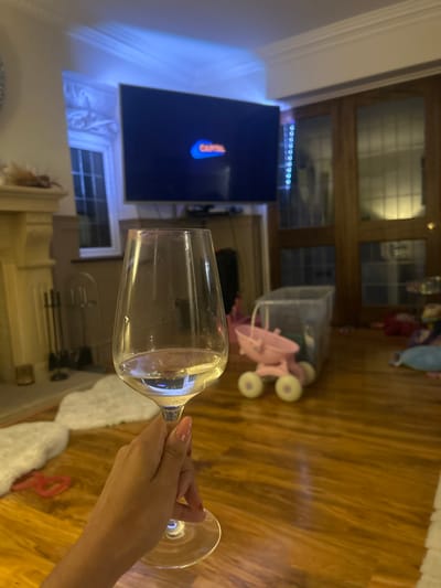 The scene is an interior residential living space located in Rushden, United Kingdom, captured under artificial lighting, consistent with evening or night. In the foreground, a right hand with light pink manicured fingernails holds a clear stemmed wine glass, approximately one-quarter full of a pale liquid. A large, wall-mounted television in the midground displays the "CAPITAL" logo. To the left, a white fireplace mantel and a window with an ornate frame are visible, accompanied by blue ambient lighting above the window and behind the TV. To the right, wooden double doors with glass panels and a vertical strip of blue LED lights are present. The floor is polished light-brown hardwood. A transparent plastic playpen or container, a pink plastic child's toy resembling a stroller, and various other scattered toys and a white fluffy rug are situated on the floor in the mid-to-background.