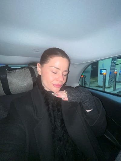 A woman with closed eyes rests in a car. She is wearing a black coat and grey fingerless gloves. Her hand is near her face, adorned with rings and silver nails. Behind her head is a grey roll with black straps. Outside the car window are multiple electronic charging stations. The car's interior is light grey.