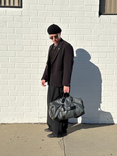 A stylish individual in a black beret, sunglasses, and a suit stands against a white brick wall on a sunny day. They carry a patterned black duffel bag. The person's shadow is cast on the wall beside them, suggesting the bright light source. The scene includes a sidewalk and the corner of a building in the background.