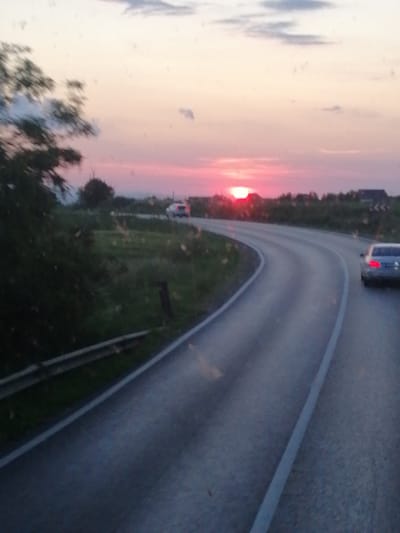 The image depicts a sunset scene on a road in Mărițeia Mică, Romania. The sun, a bright orange orb, is low on the horizon, casting a warm glow across a pale pink and orange sky. Dark silhouettes of trees and buildings line the horizon. A two-lane asphalt road curves through a landscape of green grass and scattered trees. One vehicle, a silver car, is visible on the right side of the road, its taillights illuminated. Another vehicle, a white car, is further in the distance on the left side of the road. The overall impression is of a rural setting bathed in the soft light of dusk.
