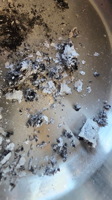 A close-up, top-down forensic image depicts fragmented ash and charred debris scattered across the reflective, scratched interior surface of a metallic vessel, likely a bowl or pan, located in Heusden, Belgium. The debris consists of irregular pieces ranging from dark black charred material to light gray and white ash, with some fragments appearing curled or crumpled. The metallic surface shows fine scratches and a semi-polished finish. No individuals, specific actions, or other environmental elements are discernible within the frame.