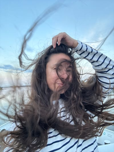 A woman with long, dark, wavy hair, which is blowing vigorously in the wind, is taking a selfie. She is wearing a white long-sleeved shirt with navy blue stripes. Her right hand is raised, holding some of her hair near her head. She appears to be on a boat, indicated by the white railing and some text on a blue and white circular emblem on the right side of the image. The emblem contains text which appears to be Cyrillic script and reads "БЕРЕГОВОЙ КОМПЛЕКС". The background is a cloudy sky and a body of water with a distant shoreline. The setting is likely the Neva River in the city of Imeni Sverdlova, Russia, a common location for boat tours and recreational activities.