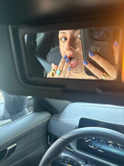 The image depicts the interior of a modern vehicle in the city of Tizapan, Mexico, captured from the passenger perspective. The primary subject is a female reflected in the vehicle's vanity mirror. She has multiple spots of light-colored cosmetic foundation or concealer applied to her face. Her mouth is open with her tongue slightly extended, displaying a blue hue. Her left hand (viewer's left), adorned with blue and white striped fingernail polish and two gold rings (one with a light blue/green oval stone), has its index finger near her lower lip. Her right hand (viewer's right), also with blue fingernail polish and a plain gold band ring, holds a black smartphone with a textured case featuring the gold initials "RV." The phone is positioned to capture her reflection. The vehicle's interior features a dark dashboard and door panel with prominent orange or tan contrasting stitching. A multi-function steering wheel is partially visible in the lower frame, and a digital instrument cluster displays a speed of 30 km/h and an odometer reading of 24374 km. A dark-colored seatbelt is visible across the woman's chest in the reflection.