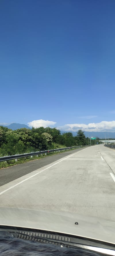 The image depicts a highway with clear blue skies and distant mountains. Lush green trees line the left side of the highway, with a metal guardrail separating the road from the vegetation. The road itself is paved concrete with white lane markings. In the distance, traffic signs are visible, indicating a junction. The foreground shows the hood of a vehicle, suggesting the photograph was taken from a moving car. The scene appears to be in Lonbaroh, Indonesia, as indicated by the presence of tropical foliage and mountainous terrain typical of the region. There are no people or animals visible in the image. The overall atmosphere is peaceful and serene, with the bright sunlight illuminating the landscape.