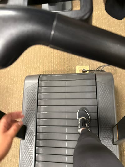 An overhead view depicts a person engaged in physical activity on a treadmill within an indoor facility in Plainsboro Township, United States. The central subject is a dark grey treadmill with a ribbed running belt. A human left foot, clad in a black athletic shoe with white accents, is positioned on the belt, while the lower left leg is encased in black athletic leggings. In the lower left quadrant, a blurred, light-skinned human right hand is visible, suggesting interaction with the treadmill's frame or controls. The treadmill rests on a textured light brown carpeted floor. Behind the treadmill, a light-colored electrical outlet is visible on the floor or baseboard, with two dark cables plugged into it, likely powering the equipment. A dark, curved support bar or handle from the treadmill's console is visible in the upper portion of the frame.