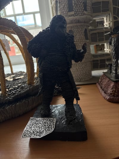 The image shows a close-up of a dark, textured figurine of a person standing on a black base. The figurine appears to be made of small, dark beads or similar material, giving it a rough, granular surface. The figure is somewhat hunched, with its arms bent, and it is holding a small, light-colored object in its right hand. The base of the figurine has a textured, rocky appearance. To the left of the figurine, part of another sculpture is visible, which seems to be composed of curved, ribbed elements, possibly representing organic forms like trees or branches. To the right of the figurine, another dark figurine with a more rigid, architectural form is partially visible, standing on a reddish-brown base.
A QR code is attached to the base of the main figurine. The background suggests an indoor setting with a window showing a bright sky and some building details. The surface on which the figurines are placed is a wooden table. The overall scene is presented in a slightly low-angle shot, with the lighting casting shadows. The location is identified as Ветрынскі сельскі Савет, Belarus.