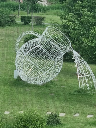 This image shows a large, white wire sculpture of a pitcher or jug with a handle and spout, situated outdoors on a grassy lawn. The sculpture is illuminated by embedded lights, giving it a luminous quality. The pitcher is the dominant subject, its intricate wireframe casting shadows on the green grass. Behind the sculpture, lush green foliage from trees forms a dense backdrop. The lawn is interspersed with rectangular, light-colored pavers, suggesting a pathway or decorative elements within the landscape. A few small, dark green bushes are visible in the mid-ground and right side of the image. The lighting suggests it could be daytime, with diffused light, possibly overcast or early morning/late afternoon. There are no people visible in the image, and no text is present. The scene appears to be a park, garden, or other outdoor public space in Kuršėnai, Lithuania, likely a decorative installation.