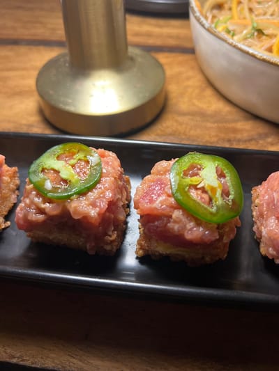 The image shows three small, square-shaped servings of tuna tartare on a black rectangular plate. Each serving is topped with a thin slice of jalapeno pepper and a small amount of what appears to be a white sauce. The plate sits on a wooden table. There is a large, golden lamp to the left of the plate, and a partially visible bowl of food is to the right. The coordinates given place this location in the city of Santa Fe, Costa Rica, but this is not visible in the image.