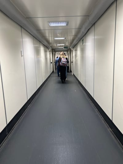 The image displays the interior of an airport jet bridge, located at Brussels Airport (BRU) in Steenokkerzeel, Belgium. The scene is an elongated, enclosed passageway characterized by white or off-white paneled walls and ceiling. The floor is a dark gray, showing signs of wear and scuff marks. Overhead, multiple rectangular fluorescent light fixtures with gridded diffusers are recessed into the ceiling, which also features a prominent gray, horizontally running insulated duct. Several individuals are observed walking away from the camera's perspective, proceeding down the jet bridge. The person closest to the camera is a female viewed from behind, wearing a dark long garment and a colorful, patterned backpack. Ahead of her, another individual, likely male, is visible in a blue top and jeans. Further down the corridor, a green 'EXIT' sign with a right-pointing arrow is affixed to the right wall. The environment is consistent with an active passenger transfer operation between a terminal and an aircraft.