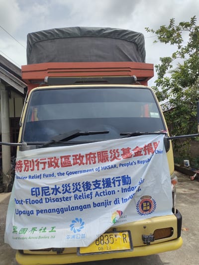 A yellow truck is parked outdoors on a paved surface, likely a street or driveway in Tanjongteungkuali, Indonesia. The truck is partially covered by a white banner with text in Chinese, English, and Indonesian, indicating a post-flood disaster relief effort sponsored by the "Special Administrative Region Government Disaster Relief Fund, the Government of HKSAR, People's Republic of China" and "Social Workers Across Borders Ltd." and "Peaceland Foundation". The banner also features logos for these organizations. The truck has a tarp-covered cargo area. The background shows a building, trees, and a cloudy sky.