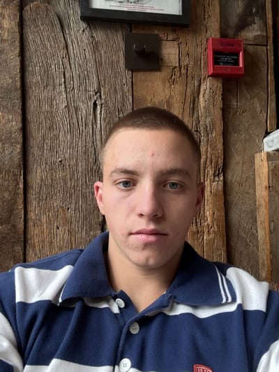 A male individual is depicted in a close-up, head-and-shoulders portrait shot indoors in Kilmersdon, United Kingdom. The subject, a young adult with light skin, short buzz-cut hair, and blue eyes, looks directly at the camera with a neutral expression. Small red blemishes are visible on his forehead and cheeks. He wears a navy blue and white horizontally striped polo shirt with a collar and three visible white buttons down the placket. A small red and white logo, possibly "KENZO", is present on the lower right chest area of the shirt. The background features a rustic, vertically paneled wooden wall. Partially visible at the top is a black-framed sign containing white text, including instructions like "DO NOT STOP TO COLLECT PERSONAL BELONGINGS." Mounted on the wooden wall are a square black electrical fixture with two circular knobs and, to its right, a red fire alarm pull station labeled "FIRE BREAK GLASS."