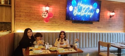 A family of three celebrates a birthday at a restaurant. A woman and two children sit at a table with plates of food, drinks, and balloons decorating the area. A TV screen displays a "Happy Birthday" message. Wall lighting, a brick-style wall, and a padded bench create a warm atmosphere.