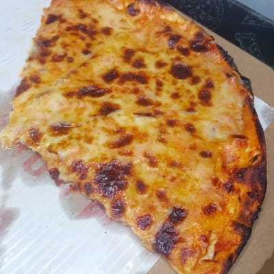 A close-up, high-angle shot shows a single slice of a cheese pizza with a golden-brown crust and bubbly, melted cheese. The pizza is placed on a white paper liner within a brown cardboard pizza box. The background is slightly blurred, but appears to be a flat surface. The scene is indoors, likely a restaurant or home kitchen, and there is no indication of El Limon, Venezuela.