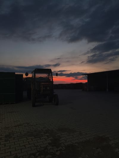 A tractor sits parked on a brick-paved lot in Galonki, Poland, at dusk. The sky above is a dramatic mix of dark, heavy clouds and a vibrant orange-red sunset, hinting at the end of a day on the farm. To the left, a stack of what appear to be dark green containers or crates stands tall. To the right, a large, dark structure, possibly a barn or warehouse, is visible. The overall scene is characterized by deep shadows and the striking contrast between the darkening landscape and the illuminated sky.