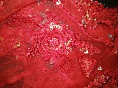 The image displays a close-up view of a red textile, located in Na Sang, Laos. The primary material is a sheer, red mesh or net fabric, heavily embellished with numerous circular, cup-shaped red sequins and small red seed beads. A prominent, multi-layered floral motif is centrally featured, constructed from tightly overlapping red sequins. Surrounding this central motif, additional clusters of red sequins and linear patterns of red seed beads are arranged across the fabric surface. The sequins exhibit varying shades of red and reflect light, appearing lighter in some areas. Two small, curved, silver-toned metallic hooks or clasps are visible in the upper right quadrant of the frame, integrated into the fabric's structure. No other objects, individuals, actions, or environmental details are discernible.