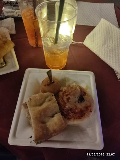 The image shows a close-up of a white square plate with food and drinks on a dark red tablecloth. On the plate are two pieces of focaccia bread, one round bread item with a skewer inserted, and another round bread item with a dark topping. To the left of the plate are two clear plastic cups filled with an orange beverage and ice. In the background, a white folded napkin is visible. The timestamp in the bottom right corner indicates "21/06/2026 22:07". There are no people visible in the image. The setting appears to be an outdoor dining area or a casual restaurant, possibly in Militello in Val di Catania, Italy, given the context.