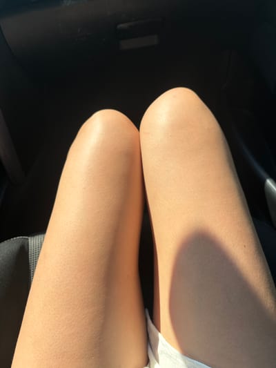 The image shows a close-up of a person's bare legs, with the skin illuminated by sunlight casting a shadow across the upper thigh. The setting appears to be indoors, possibly a car interior, indicated by the dark, textured surface on the left. The person is sitting, with their knees bent. A piece of white fabric is visible on the right side of the frame, possibly clothing or a seat cover. The skin texture is visible, with small bumps, and there is a slight sheen from the light. No text is visible in the image. The scene suggests a casual moment, possibly during daytime given the strong sunlight.