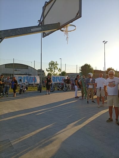 An outdoor, low-angle, full shot captures a group of people standing near several motorcycles on a paved surface under a clear, bright sky. A basketball hoop with a net hangs in the foreground on the left, casting a long shadow across the ground. In the background, a wire fence encloses an area with a building and trees. A banner with text in Italian, including "Arbia," is visible on the building. The people are dressed in casual attire, and some are holding drinks or talking. The lighting suggests it is late afternoon or early evening.
