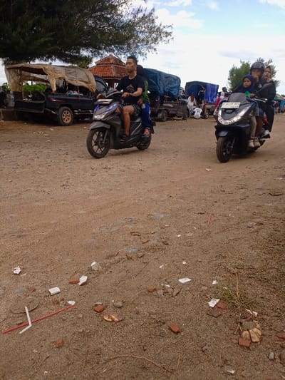The image depicts a scene in Pameungpeuk, Indonesia, located at -7.668 latitude and 107.694 longitude. The scene features a sandy, dirt road, two motorbikes, and several people.  A man, wearing a blue shirt and black shorts, is riding a motorcycle with a pillion passenger. Behind him, two women and a child are riding another motorcycle. The two motorcycles are in motion, traveling in the same direction, and the road is empty in both directions. There is a large, covered flatbed truck parked on the left side of the road. The scene is a clear day with a partly cloudy sky.