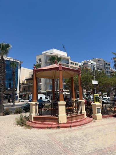 The image shows a bright, sunny day in Gżira, Malta, with a clear blue sky. In the foreground, a red and yellow gazebo with wooden pillars stands on a paved area. Inside the gazebo, two individuals are seated. To the left of the gazebo, a palm tree is visible. In the background, several multi-story buildings line a street with palm trees planted along the sidewalk. A white van and other vehicles are present on the street. A lamp post with two globes stands next to the gazebo, and a yellow parking meter is also visible. The overall scene depicts an urban environment with a mix of modern architecture and natural elements.