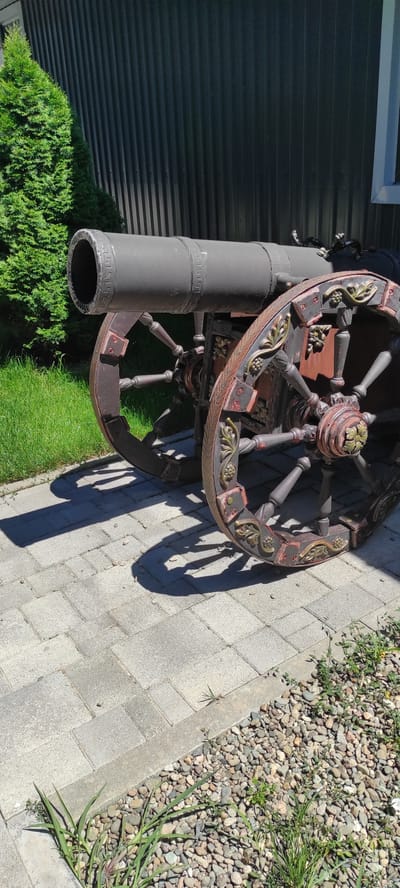 An antique cannon is positioned outdoors on a paved walkway, with a green coniferous tree and a dark, ribbed wall in the background. The cannon has a dark grey barrel and intricately decorated wooden wheels with red and gold accents. The ground is covered with grey rectangular pavers, and a section of small pebbles is visible to the right. Shadows of the cannon and the tree are cast on the paved area. The scene appears to be set in a garden or courtyard.