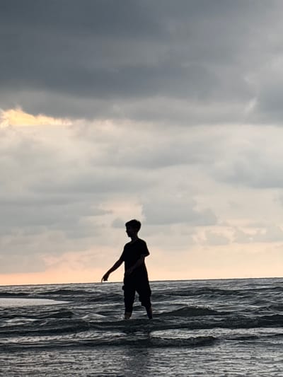 A silhouette of a person stands in shallow water at the beach, likely in Angkleng Barat, Indonesia. The sky is overcast with dark clouds above and lighter, sunlit clouds near the horizon. The water appears rippled and the image is oriented vertically.